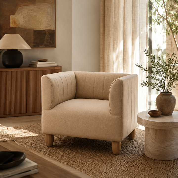 A beige upholstered accent chair with rounded arms and wooden legs, placed in a sunlit living room beside a small round side table and a wood cabinet.